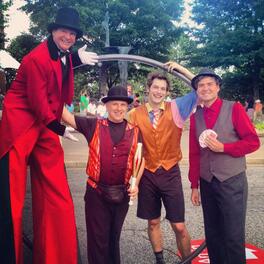 Stilt Walker, Juggler with club, Acrobat with metal wheel, and Magician with playing cards posing outdoors at a festival. 