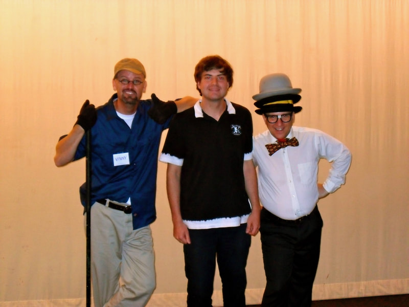 Actor dressed as Janitor with broom and Juggler with 3 hats and a bow tie pose with producer Thomas Seaton in front of a white theatre curtain. 